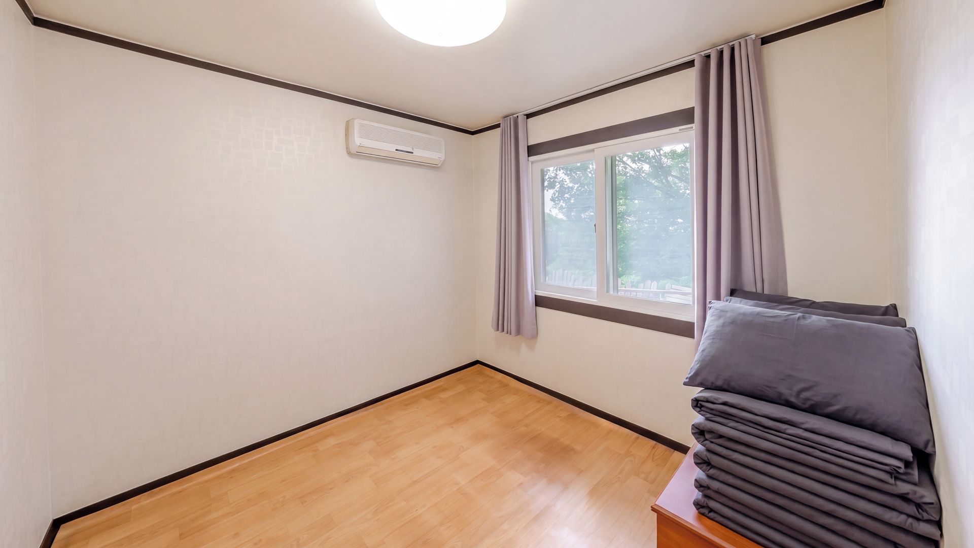 Una habitación con piso de madera, una ventana con cortinas y un montón de mantas dobladas sobre una mesita de noche de madera.