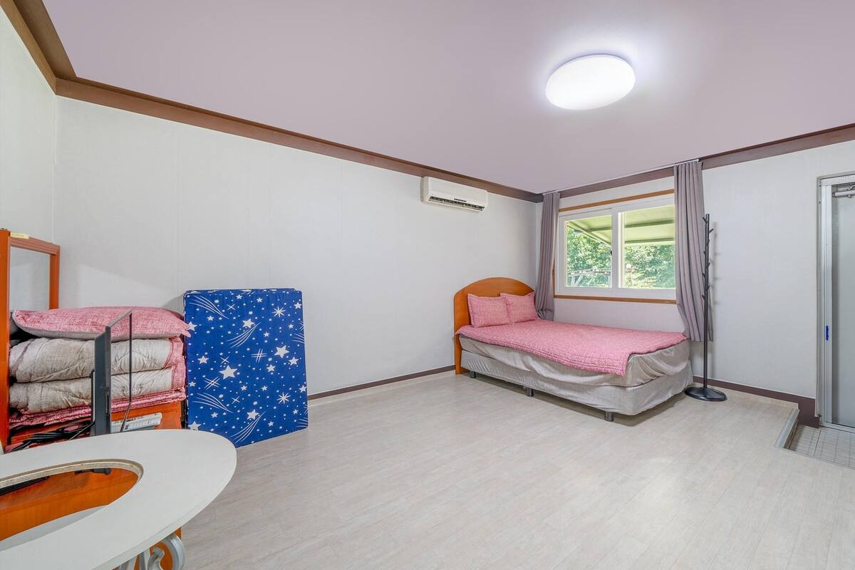 Ein kleiner, gut beleuchteter Raum mit einem Einzelbett, einem Fenster mit Vorhängen, einem Heizkörper und einer Tür.
