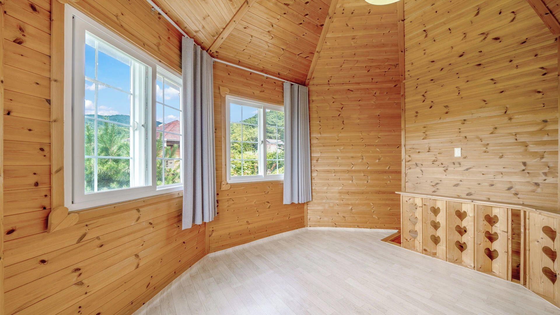 A room with wooden walls and floors, large windows with curtains, and a view of greenery outside.