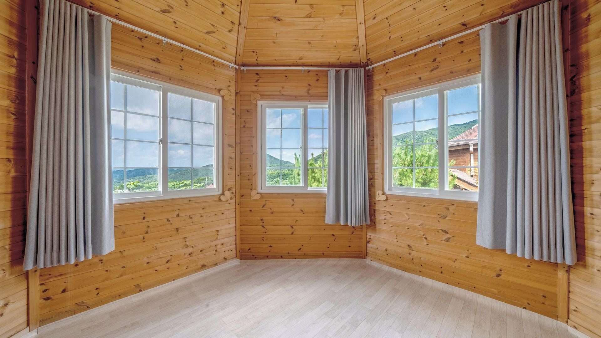 A room with wooden walls and floors, large windows with curtains, and a view of greenery outside.