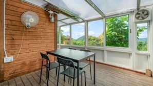 Una terraza de madera con mesa y sillas, amplias ventanas con vistas a zonas verdes y un ventilador en la pared.