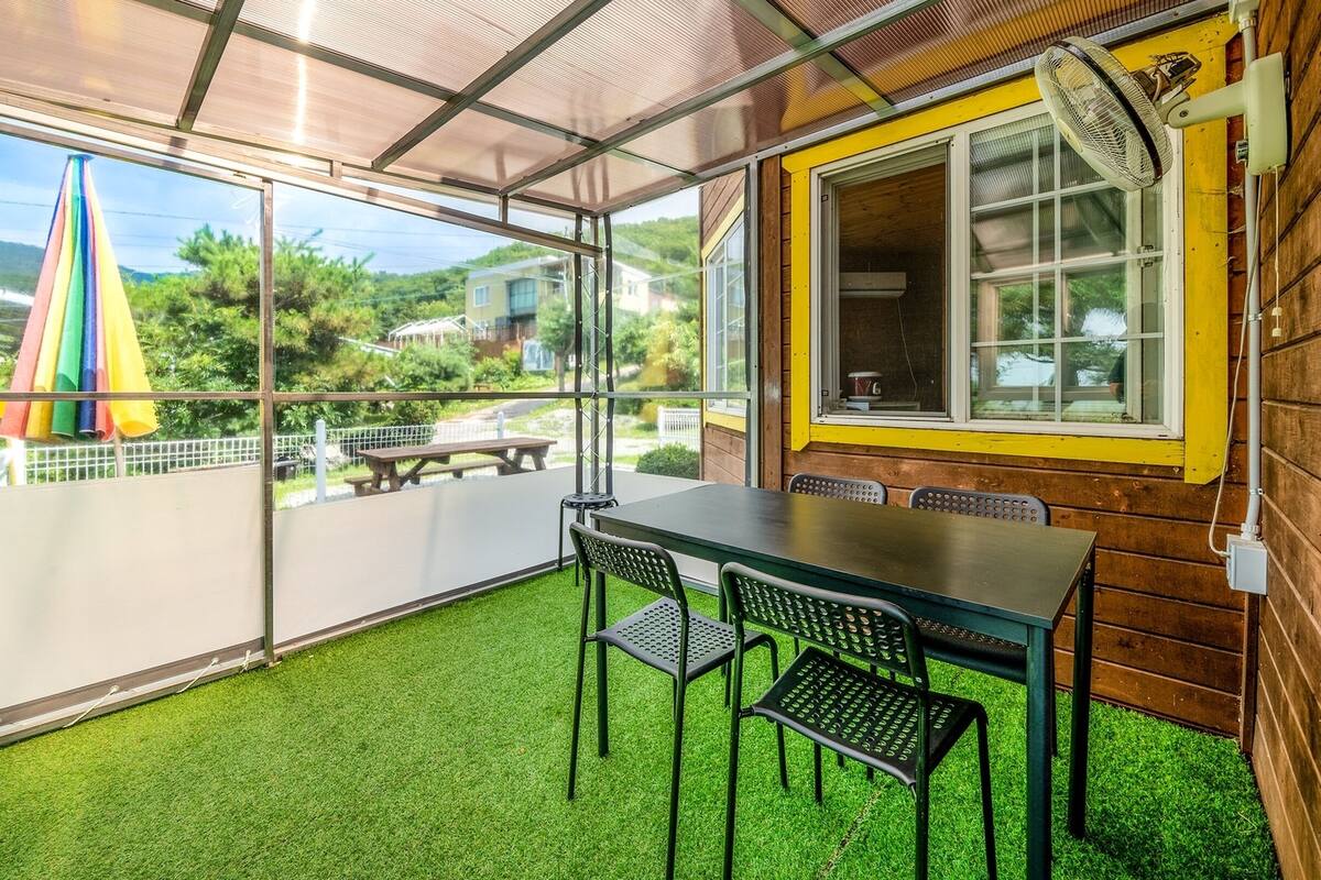 Eine überdachte Terrasse mit Tisch und Stühlen, Blick auf eine grüne Landschaft und ein Fenster mit Ventilator.