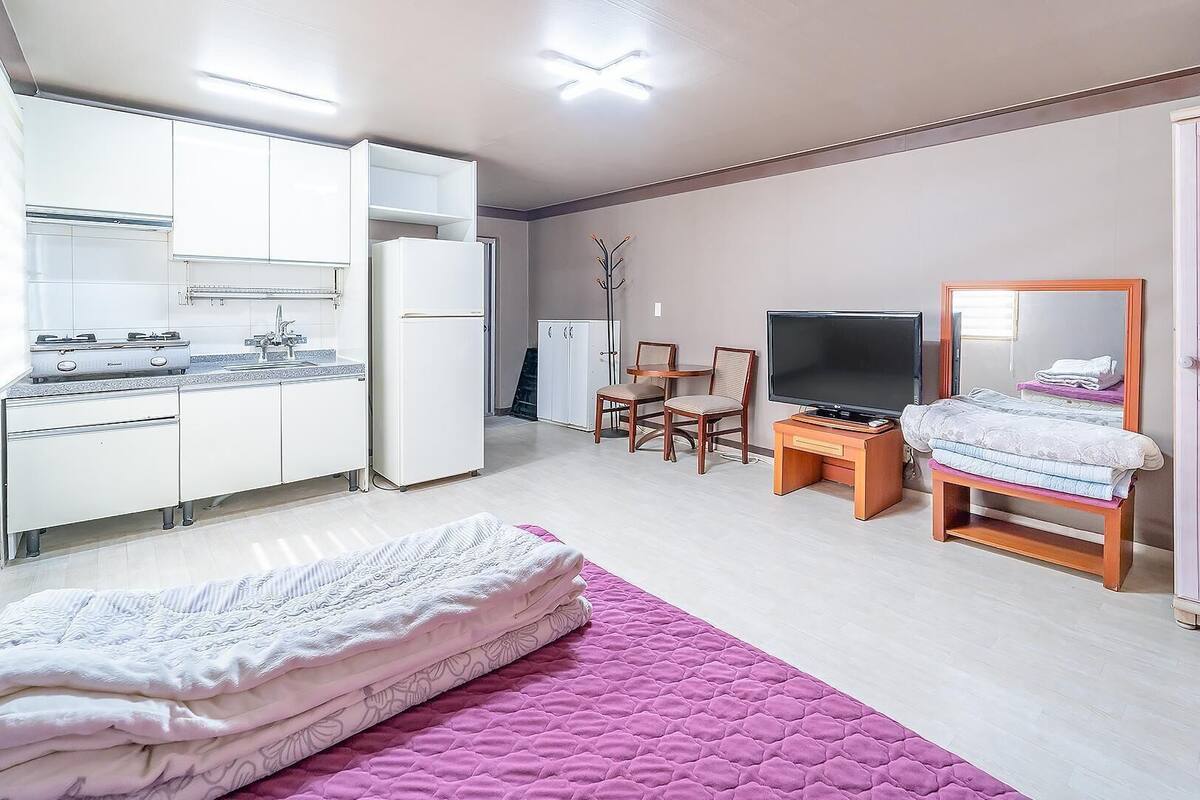 Ein gemütlicher Raum mit Küchenzeile, einem Bett mit rosa Bettdecke, einem Fernseher und einem Kleiderschrank.