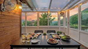 A covered dining area with a table set for a meal, large windows with a view of greenery, and a ceiling fan. - Paju Gamaksan Sanchon Village Pension (Paju)