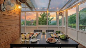 Un área de comedor techada con una mesa lista para una comida, ventanas amplias con vista a follaje y un ventilador de techo.