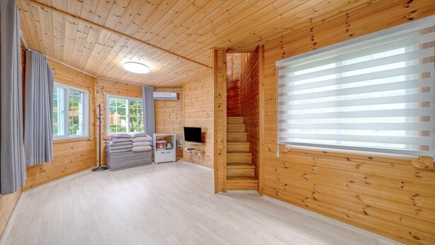 Interior de una cabaña de troncos con piso de madera, chimenea, televisor y ventana con persianas.