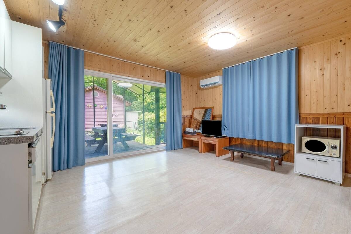 Ein Zimmer mit Holzboden, einer großen Glasschiebetür, die nach draußen führt, einer Holzbank, einem Fernseher und einem Radio.