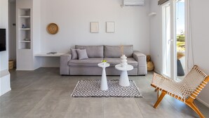 Een moderne woonkamer met een grijze bank, twee witte bijzettafels, een geweven fauteuil en een kamerkleed met een patroon.