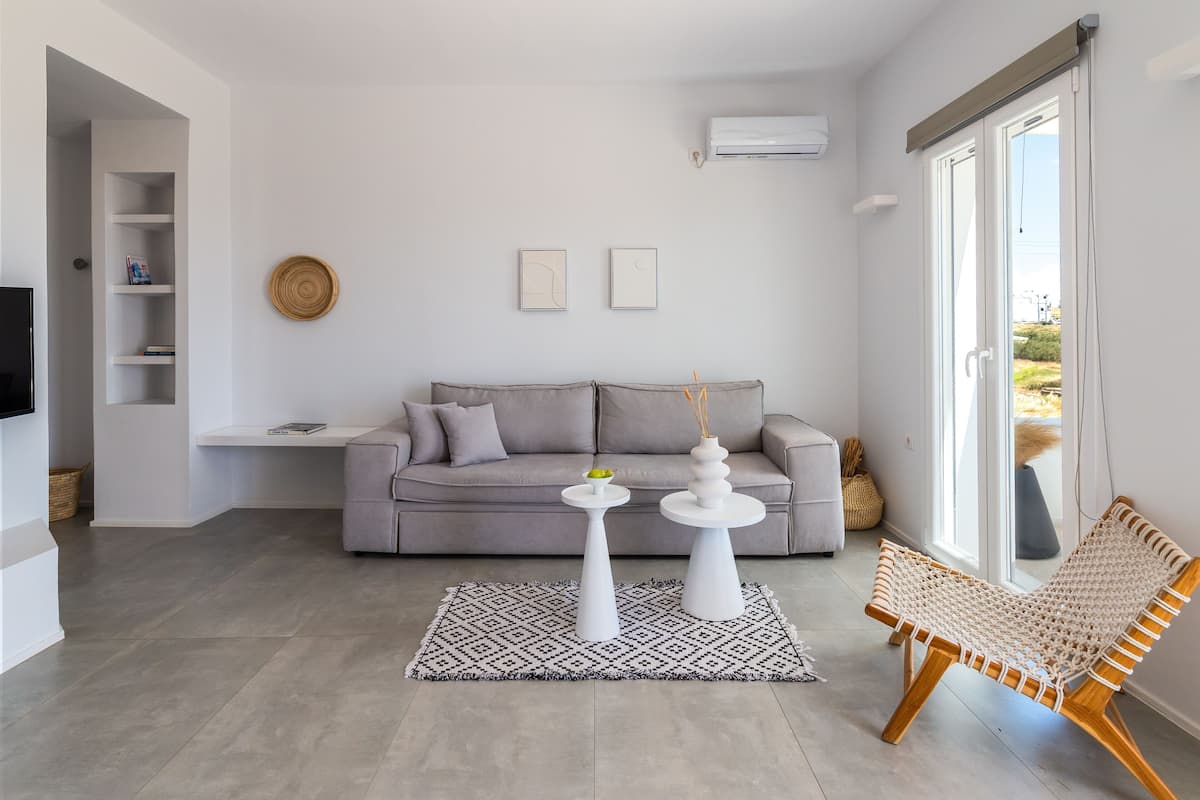 Een moderne woonkamer met een grijze bank, twee witte bijzettafels, een geweven fauteuil en een kamerkleed met een patroon.