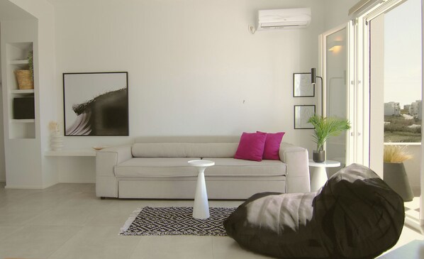 A modern living room with a beige sofa, a white coffee table, and a black bean bag. There is a framed picture on the wall and a potted plant on a side table. - AELIA Tinos (Tinos)