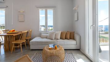 Una sala de estar moderna con un sofá blanco, sillas de madera, una mesa de comedor con fruta y vistas de la ciudad a través de grandes ventanales.