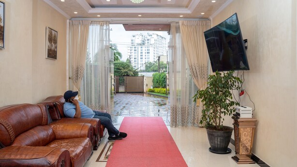 Un vestíbulo de hotel con alfombra roja, sofá de cuero, una planta grande en maceta, televisor de pantalla plana y vista a un edificio a través de una puerta de vidrio.