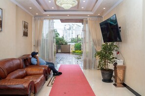 Eine Hotelhalle mit rotem Teppich, einer Ledersofa, einer großen Pflanze im Topf, einem Flachbildfernseher und Blick auf ein Gebäude durch eine Glastür.