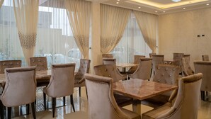Un comedor con varias mesas y sillas, amplias ventanas con cortinas y un candelabro.