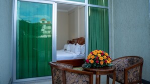 Habitación de hotel con un ventanal grande, una cama, una silla de madera y una mesa con un arreglo floral.