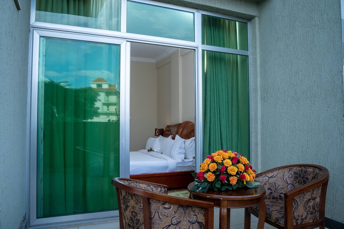 A hotel room with a large window, a bed, a wooden chair, and a table with a flower arrangement.