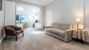 Un salon avec un fauteuil en cuir marron, un canapé beige et une petite table sur laquelle se trouve une lampe. Au fond, on aperçoit un coin cuisine avec une porte blanche et une fenêtre équipée de stores.
