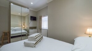 Un lit bien fait, avec une pile de serviettes pliées, une table de chevet avec une lampe, un miroir et une fenêtre avec des stores.