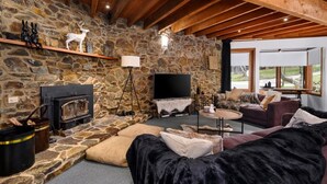 Ein gemütliches Wohnzimmer mit Steinwand, Holzbalken, Kamin, Flachbildfernseher und einer bequemen Sitzecke mit Blick ins Freie.