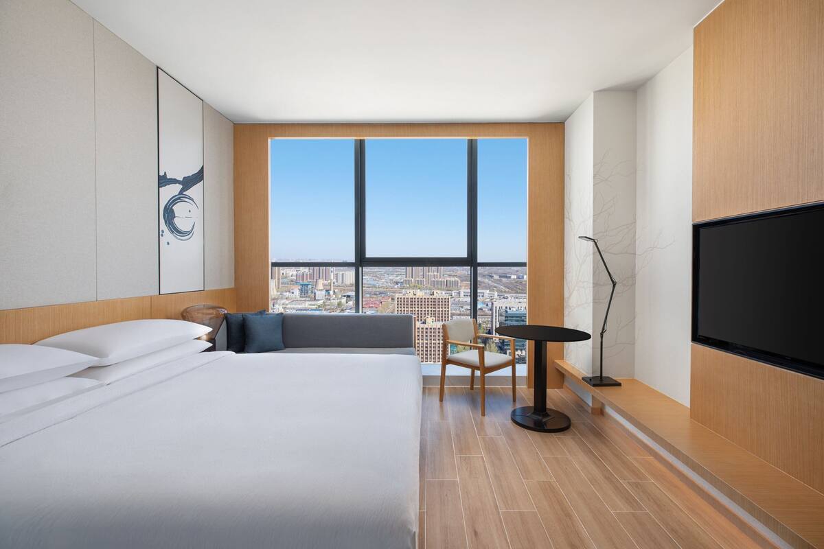 A modern hotel room with a large bed, a city view, a desk, and a TV.