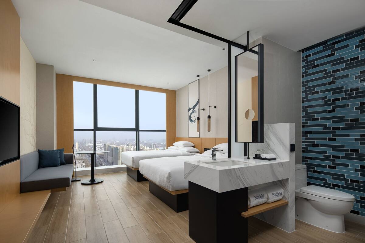 A modern hotel room with a large window, two beds, a bathroom with a marble sink, and a city view.