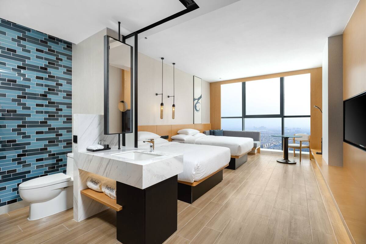 A modern hotel room with a large bed, a separate sofa, a bathroom with a marble sink, and a city view through large windows.