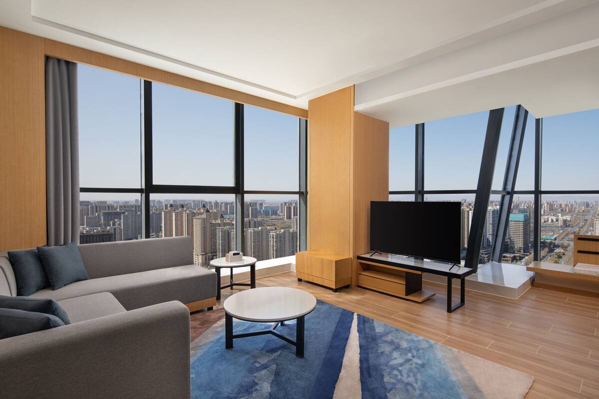 A modern living room with a large TV, a sofa, a coffee table, and a view of the cityscape through large windows.