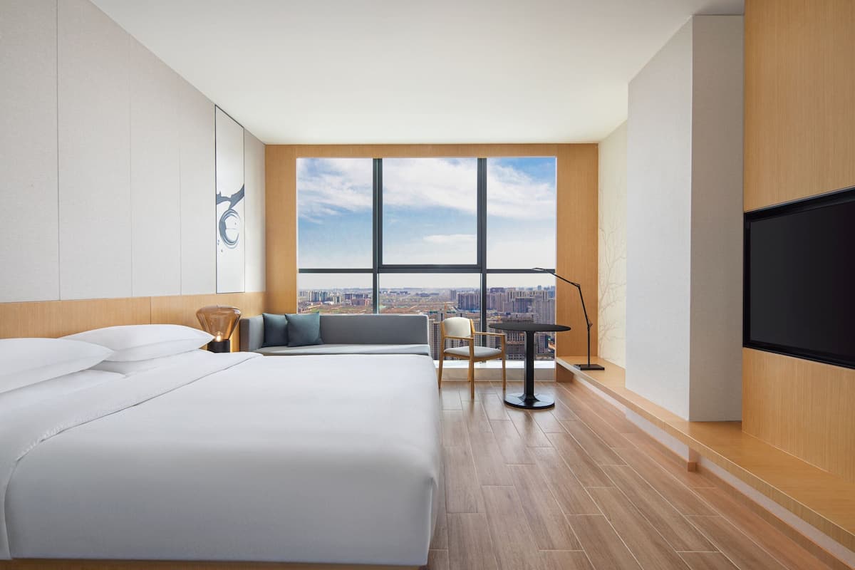 A modern hotel room with a large bed, a seating area with a table and chairs, a large window with a city view, and a flat-screen TV mounted on the wall.