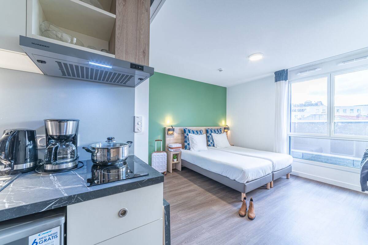 A modern hotel room with a bed, a small kitchenette, a window with a city view, and a wooden ceiling.