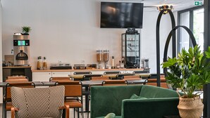 Un café moderno con una televisión de pantalla plana, máquinas de café y diversas opciones de asientos, incluyendo sofás verdes y sillas estampadas.
