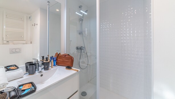 Un baño moderno con ducha de vidrio, un lavamanos blanco y diversos artículos de aseo, y un espejo sobre el lavamanos.