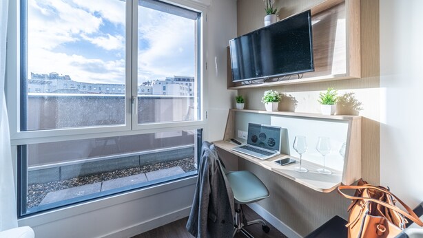 Una habitación moderna con un ventanal grande, un escritorio con una computadora portátil, un televisor de pantalla plana y una pequeña planta en maceta.