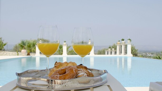 A dining table with a tray of pastries and two glasses of orange juice, overlooking a pool with a view of the sea.