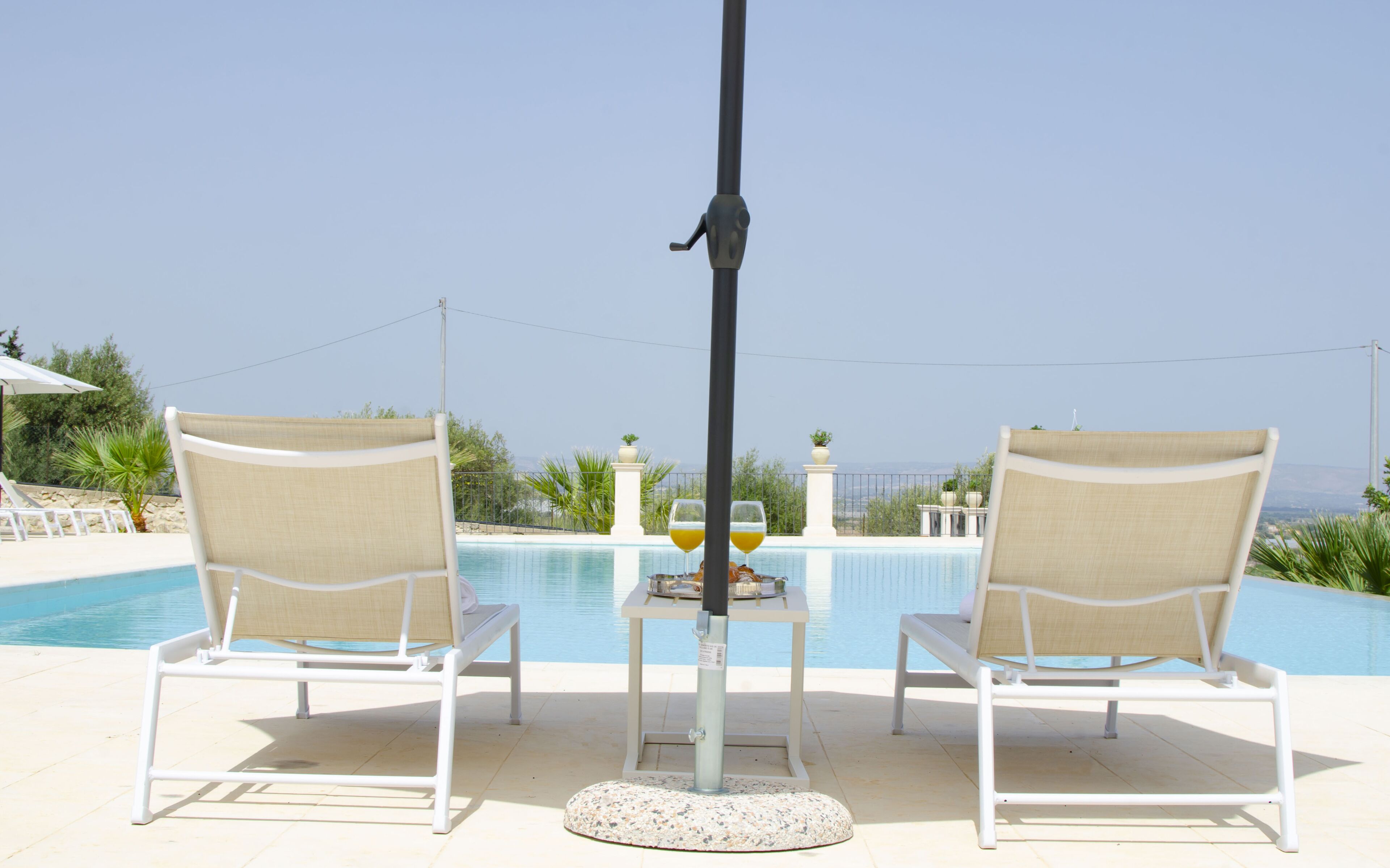 Two white lounge chairs with beige cushions facing a pool, a table with two glasses of orange juice, and a clear view of the sea and mountains in the background.