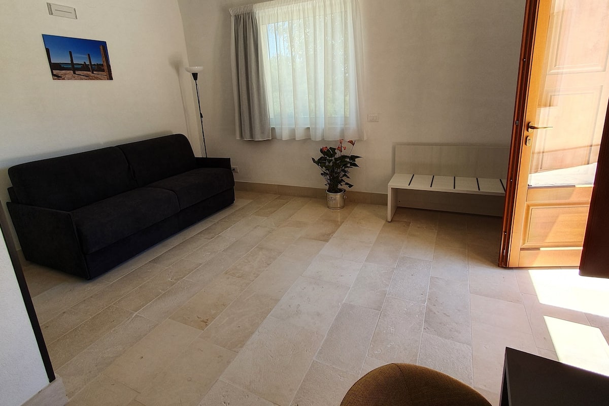 Una sala de estar con un sofá negro, un banco blanco, una planta en maceta y una ventana con cortinas.