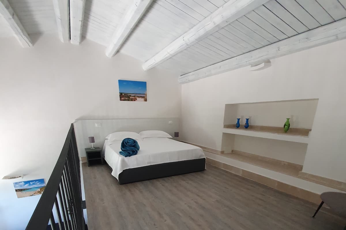 Un dormitorio con una cama, una mesita de noche, una pequeña mesa, una silla, una estantería con adornos y un cuadro en la pared.