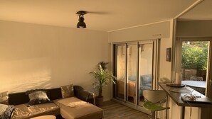 Un salon moderne avec un canapé, un pouf et une petite table ronde. On y trouve une plante en pot et une porte-fenêtre coulissante donnant sur un balcon.