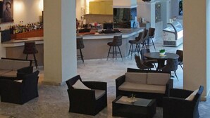 Amplia sala de estar con diversas opciones de asientos, incluyendo sofás y sillones, una zona de bar con botellas, y una cocina al fondo.