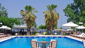 Una piscina con sillas de descanso y sombrillas, rodeada de palmeras y una zona cubierta con mesas y sillas.