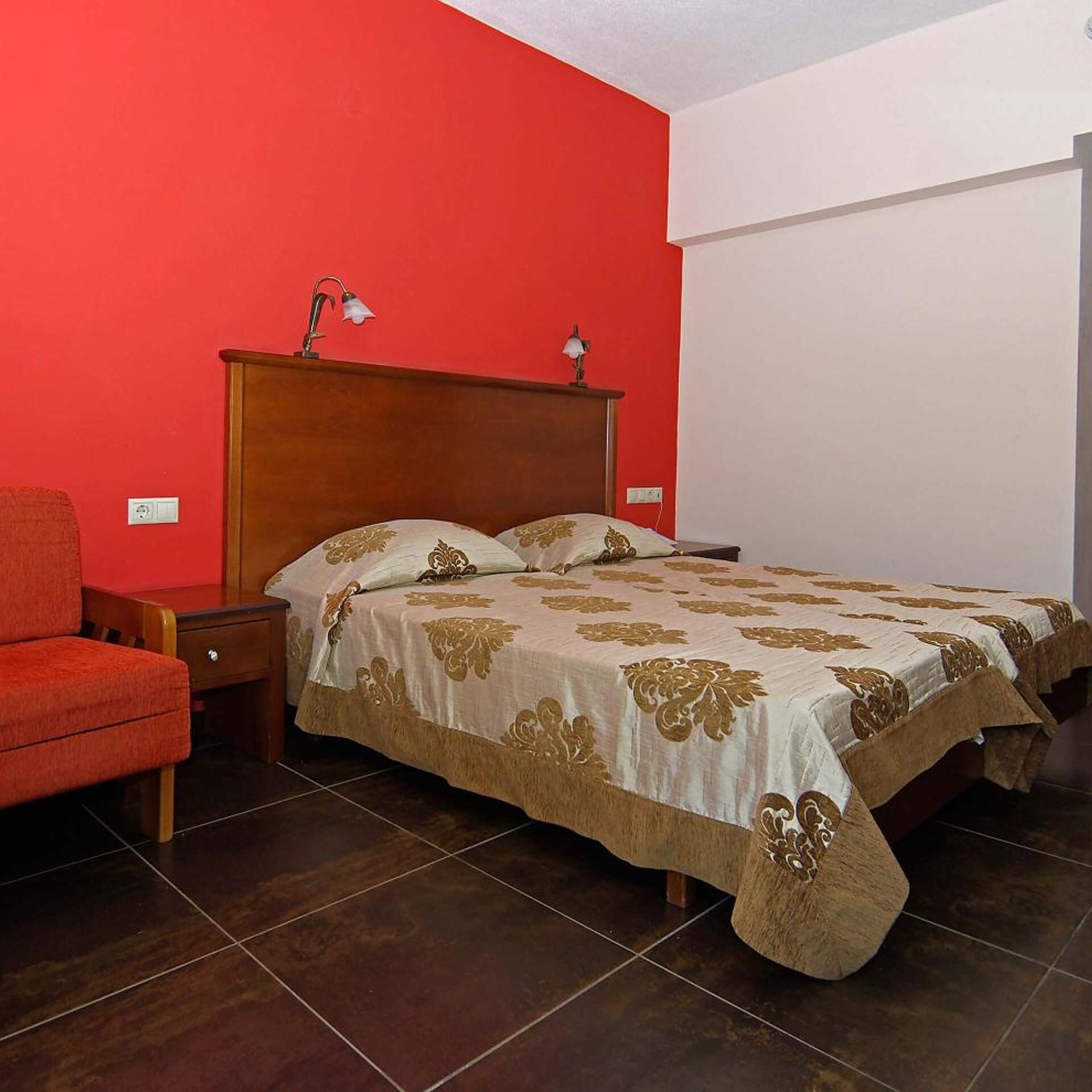 Een slaapkamer met een rode wand, een houten hoofdbord, een bed met een beddengoed met een patroon, een nachtkastje en een rode fauteuil.