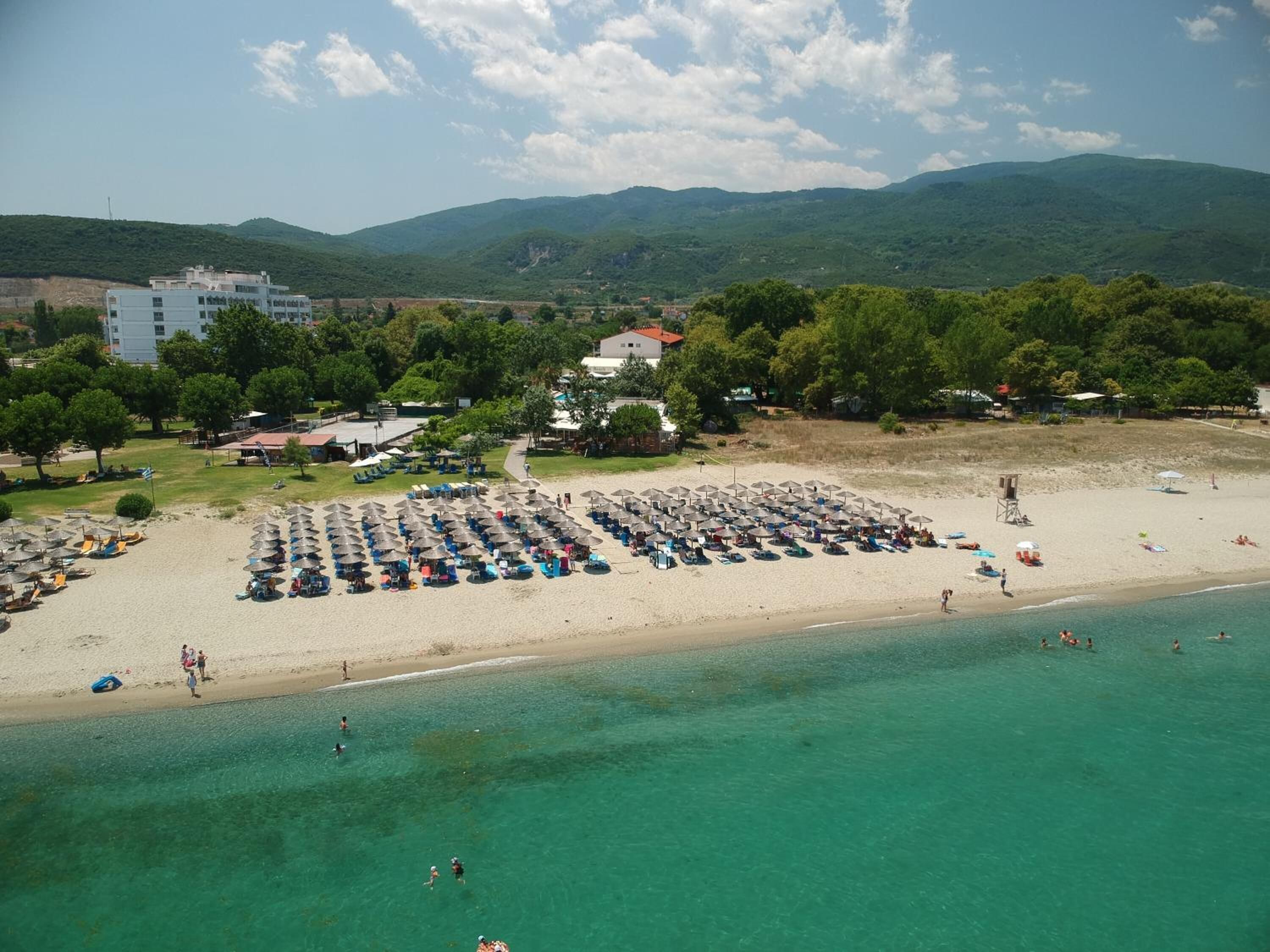 A beach with numerous umbrellas and sun loungers, a clear blue sea, and buildings in the background.