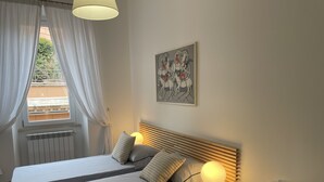 Une chambre à coucher comprenant un lit, une lampe de chevet, une chaise, une fenêtre avec des rideaux et un tableau accroché au mur.