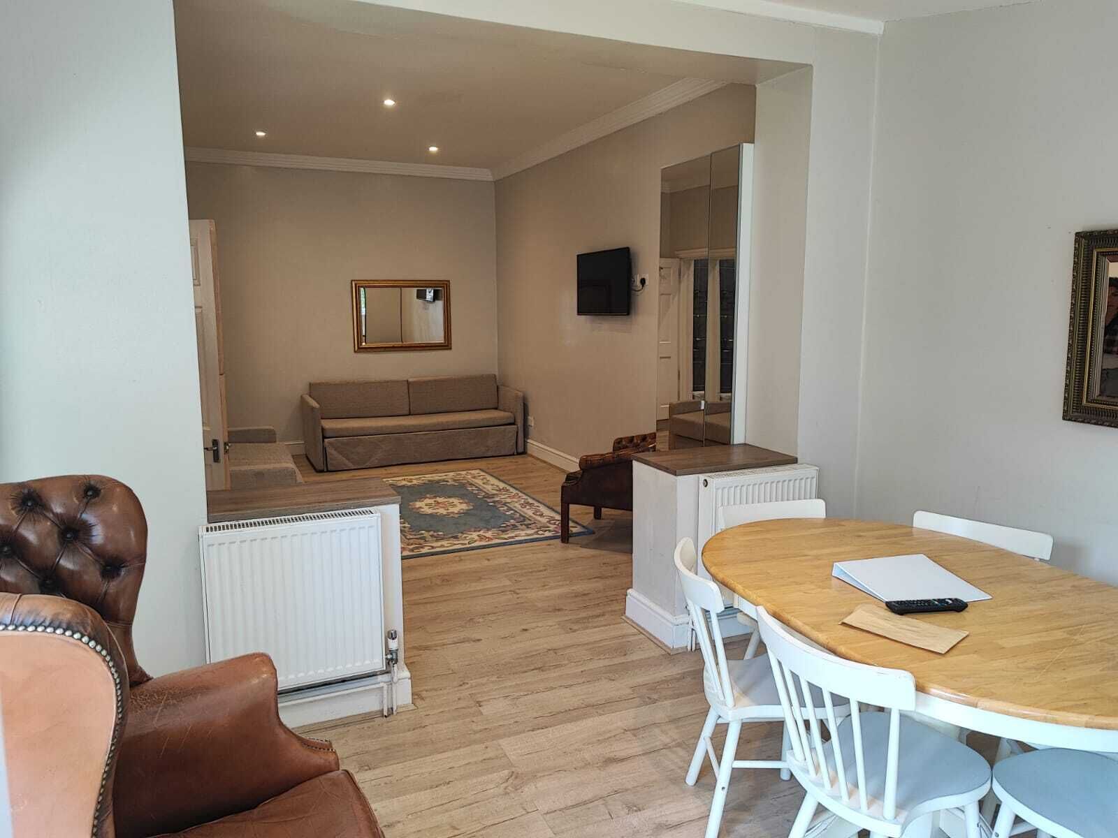 A living room with a sofa, a coffee table, and a dining table with chairs. There is a radiator, a mirror, and a television on the wall.