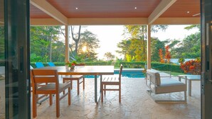 A covered outdoor dining area with a pool view, stone flooring, and wooden furniture.