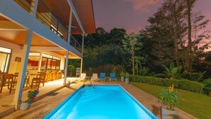 A modern two-story house with a swimming pool, outdoor seating, and lush greenery.