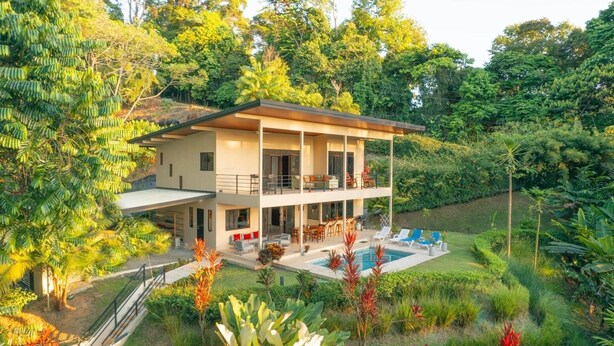 A two-story house with a balcony, a swimming pool, and lush greenery surrounding the property.