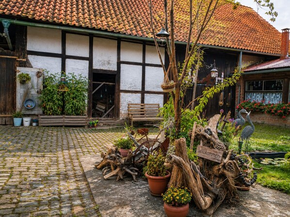 A courtyard with a cobblestone path, wooden benches, potted plants, and a rustic sign.