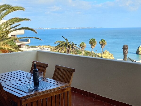 A wooden table with two chairs on a balcony overlooking a coastal view with palm trees and a clear blue sea. - Ocean View • Walk to Beach • Pool • Lagos • 64 (Lagos)