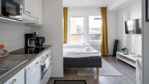 A modern kitchen with a built-in microwave, coffee maker, and oven. There is a bed with white linens and a bedside table with a bowl and a plant.