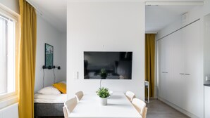 A modern, well-lit room with a dining table, chairs, a bed with a yellow pillow, a mounted TV, and a potted plant.